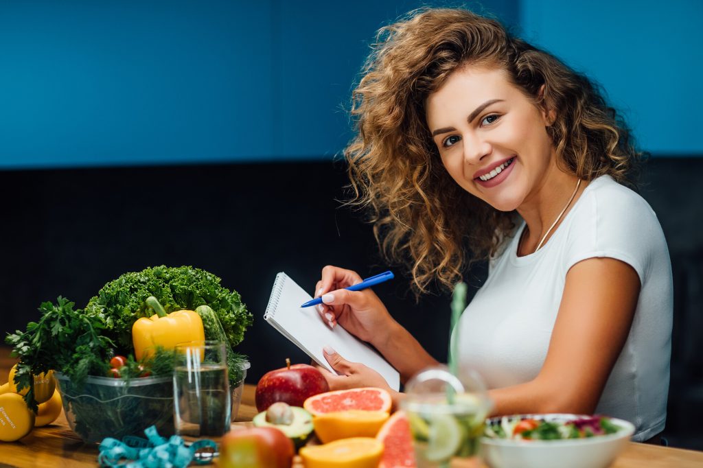 ung kvinna sitter med block och penna vid ett bod med grönsaker och frukt  som passar för pegan-dieten