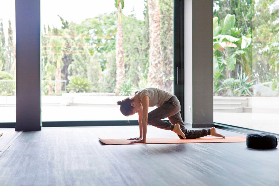 kvinna praktiserar yoga övningar i en motionssal med stora fönster, palmträd i bakgrunden