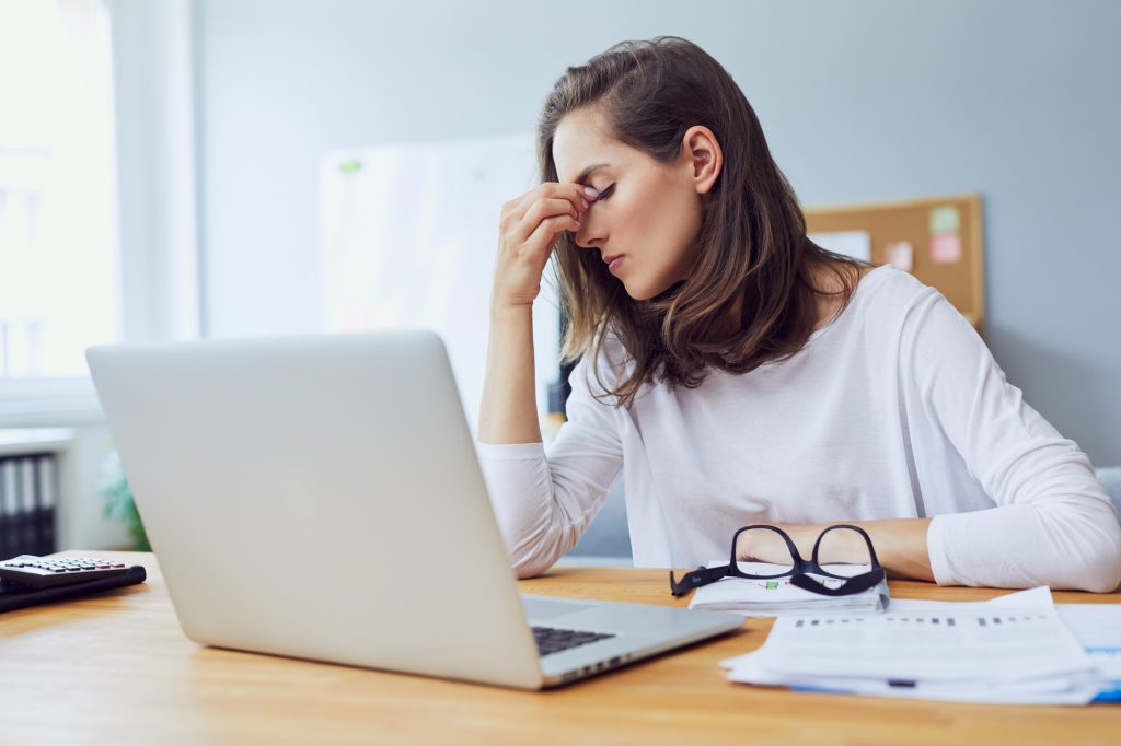 En ung kvinna sitter framför datorn, ser stressad ut och håller sig för huvudet pga smärta.